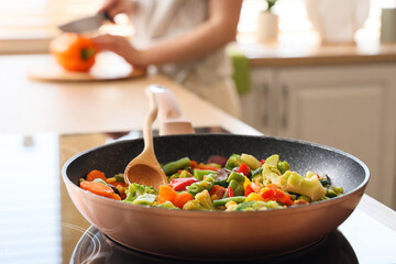 Frying pan with vegetables on stove against woman cutting bell pepper in kitchen, closeup