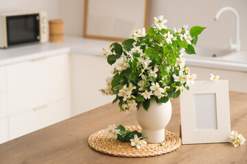 Vase of blooming jasmine flowers with blank frame on table in kitchen