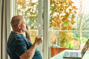 Middle-aged man with laptop, drinking coffee, talking on phone, enjoying window view.