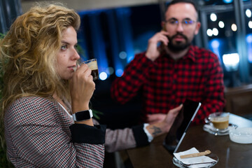 Business meeting in a cozy cafe setting with two young professionals enjoying espresso and engaging in conversation. Woman wearing smartwatch sipping coffee while man in plaid shirt uses smartphone.