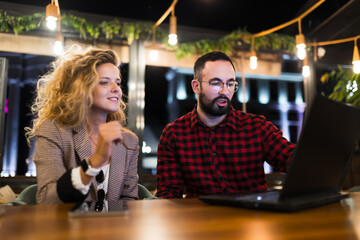 Two colleagues collaborating in a modern office space during the evening, discussing ideas and sharing insights at a laptop while sitting at a wooden table, surrounded by stylish decor and ambient