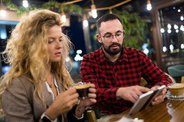 Young man and woman enjoying coffee while discussing with a tablet at a cozy cafe. Casual meeting and engaging conversation in a warm, inviting coffee shop setting during an evening out.