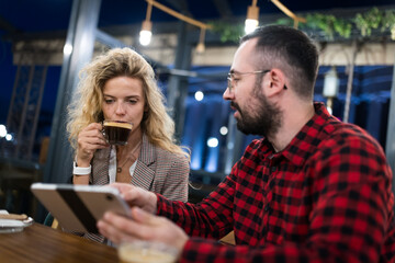 Two young adults in a cozy coffee shop engage in conversation while sharing a digital tablet, creating a warm and inviting atmosphere perfect for casual meetings or friendly gatherings.