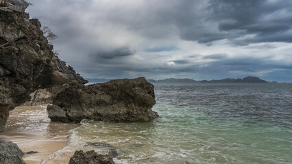 Picturesque boulders in the ocean. Coastal cliffs overhang the water. Sea waves are foaming on a sandy beach. There are no people. Mountains on the horizon. Clouds in the sky. Philippines. Palawan