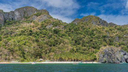 Beautiful tropical sandy beach. View from the ocean. Boats near the shore. Picturesque cliffs. Silhouettes of people relaxing on the white sand. Lush green vegetation on the mountainside.Blue sky