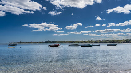 Fishing boats, motorboats are anchored in the ocean. Crystal clear calm water. Green vegetation on the shore in the distance. Blue sky, clouds. Mauritius.