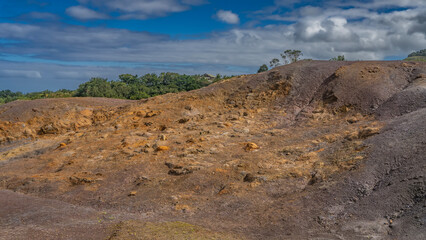 A unique geological landmark. 23 coloured earth of Mauritius. Multi-colored sand hill against a background of blue sky and clouds. Green vegetation in the distance. La Vallée Des Couleurs Nature Park
