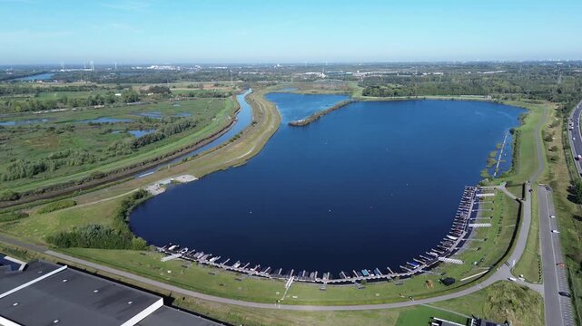 Drone flies north over the lake. Piers with boats, the Dijle River and lush nature are visible. In the distance, several wind turbines rise above the landscape near Mechelen, Belgium.