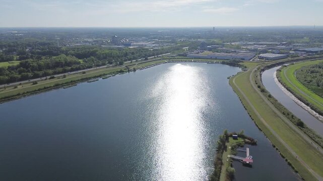 Drone flies southeast over the lake towards Mechelen North. Sunlight sparkles on the water. Visible: Dijle River, E19 highway, industrial area and green landscape.