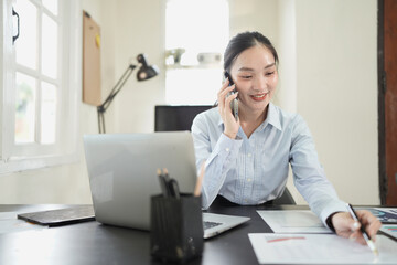 Obraz premium Asian businesswoman talking on cell phone while analyzing documents and working on laptop in office.