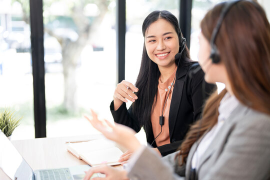 Two professional Asian call center agents collaborate, smiling and wearing headsets in a modern office.