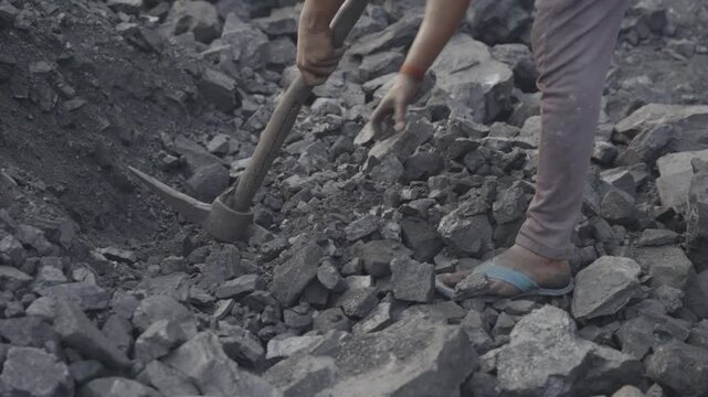 Young male worker extracting coal at coal mine, wearing open toed sandal and pant, the individual is actively engaged in breaking or moving jagged pieces of coal using a hand tool, Bokaro, Jharkhand