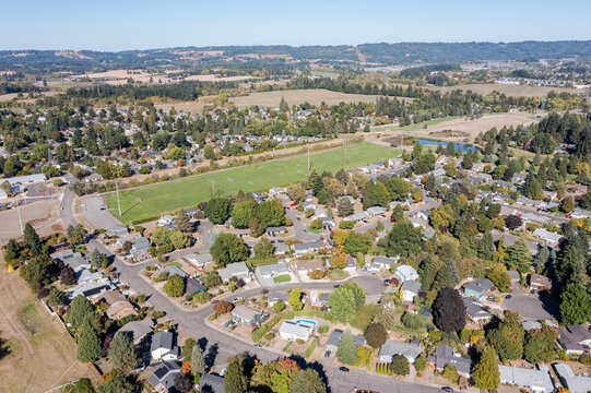 4K aerial drone photo of a suburban residential neighborhood in Hillsboro Oregon with tree lined streets, houses and a large open green field with sports fields surrounded by rural landscape and hills