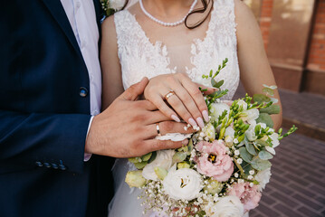 The bride and groom embrace each other on their wedding day and hold a bouquet of flowers on which they have placed their hands and are showing off their gold wedding rings