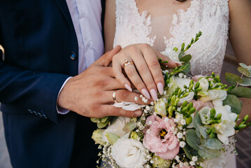 The bride and groom embrace each other on their wedding day and hold a bouquet of flowers on which they have placed their hands and are showing off their wedding rings