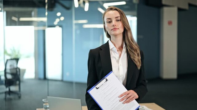 Focused businesswoman, dressed in a black blazer and white shirt, confidently poses with a clipboard in a modern office environment. Her serious expression conveys determination and readiness for work