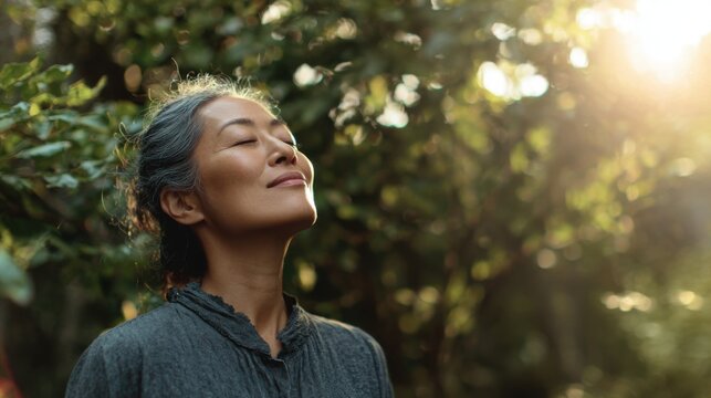 Medium view of a healthy Asian adult outdoors, standing in nature, closing eyes while taking a deep breath of fresh air, smiling with a calm and energized expression, posture open and relaxed
