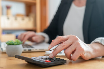 Person's hand operating a calculator while typing on a laptop, performing financial calculations, accounting tasks, managing business reports, and analyzing expenses in an office setting