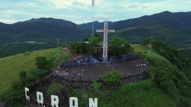 Aerial view of Mount Tapyas summit with white cross and CORON letters on a green hill above forested ridges under a cloudy evening sky