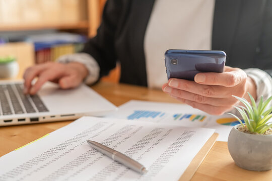 Businesswoman's hands are multitasking, typing on a laptop and holding a smartphone while reviewing financial data, charts, and graphs on printed reports at a wooden desk