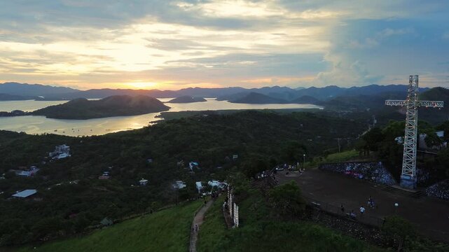 Sunset view from Mount Tapyas shows the hilltop cross above Coron Bay with island silhouettes and glowing water beyond the green hillside
