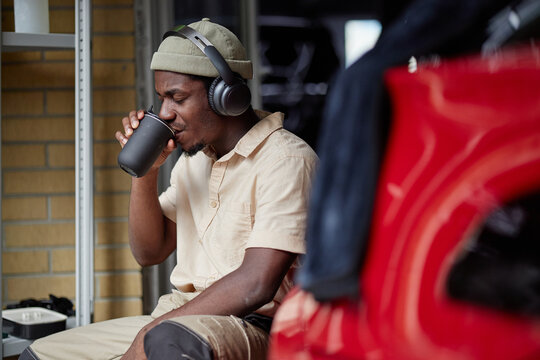 Young adult Black man wearing headphones sitting near red car, drinking from cup, eyes closed, appearing relaxed, short beard visible, brick wall and metal shelves in background