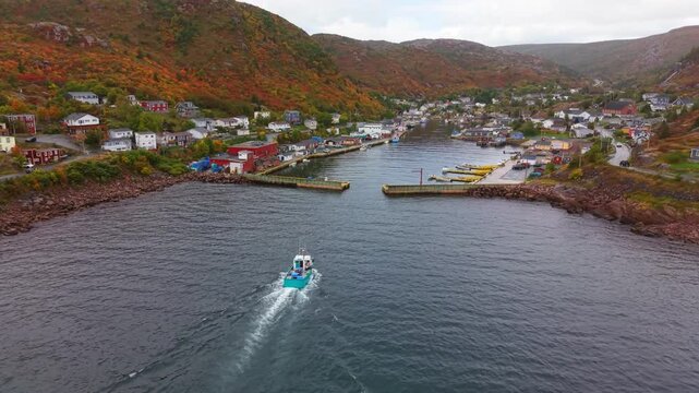 High drone shot over Petty Harbour, Newfoundland, in autumn captures a boat leaving the harbor, its wake trailing across calm waters, framed by rugged hills and colorful fall foliage.