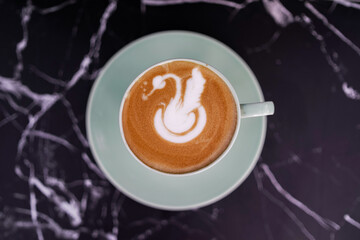 Top view of a cup of cappuccino with swan latte art served on a green ceramic saucer over a marble table, highlighting elegant coffee presentation.