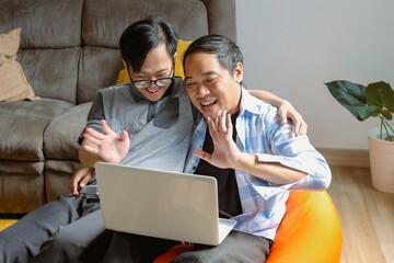 Asian Father and Son Sitting on Bean Bag and Using Laptop for Video Calling at Home