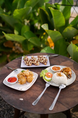 Top view of assorted Indonesian dishes including fried chicken with rice, almond croissant dessert, and noodle ball appetizer served on a rustic wooden table outdoors.