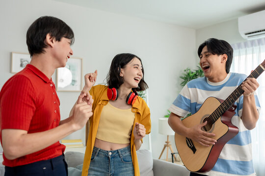 Diverse Asian friends playing guitar and dancing in living room at home. 