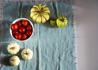 op view of ripe red cherry tomatoes in a white bowl alongside assorted gourds placed artistically on a textured blue fabric. The image highlights autumn harvest and organic produce.