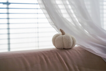 Cozy white fabric pumpkin rests on pastel sofa beside flowing curtain, with blurred window blinds behind, creating a soft minimal autumn home decor scene with gentle natural light. Copy space