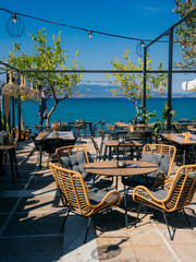 Sunny outdoor terrace of a seafront café in, Central Greece. Elegant wicker chairs, tables, and large cream umbrellas create a relaxing Mediterranean atmosphere with stunning views of the turquoise 