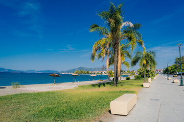 Sunny view of the seaside promenade in Kamena Vourla, a coastal town in Central Greece. The photo shows palm trees, benches, parasols, and the calm blue waters of the North Euboean Gulf under a clear