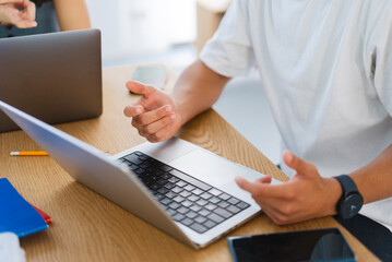 Two people are collaborating on laptops at a wooden table. One person gestures with their hands while discussing work, with a smartphone and smartwatch nearby.