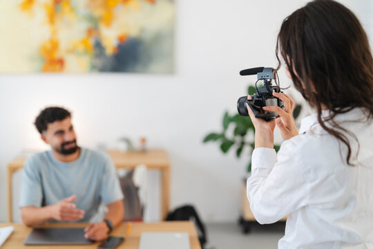 A woman films a man with a professional camera setup, including an external microphone. The man sits at a desk, gesturing as he speaks, creating video content.