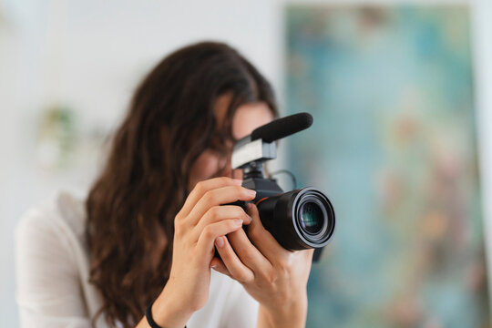 A woman with long, dark hair holds a mirrorless camera equipped with an external microphone. She is focused on recording video or capturing photographs for her creative project.