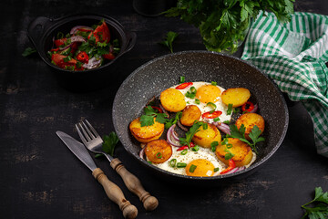 Fried eggs and potatoes with onions and chili peppers and fresh salad in a  dark background.