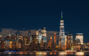 Manhattan skyline of New York City at night with glowing skyscrapers and reflections on water,...