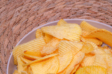 Crispy golden potato chips stacked in a white bowl on a woven mat. The crunchy texture and warm...