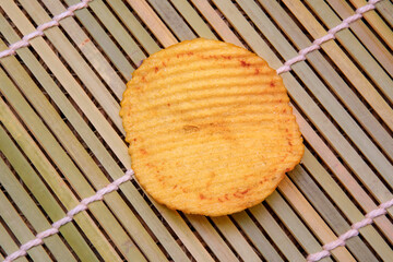 A single ridged potato chip placed on a bamboo mat. The golden-yellow crisp shows a crunchy texture with slight browning around the edges. Highlighting its salty and appetizing snack appeal.