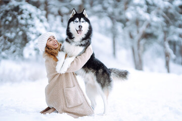 A young woman in a warm coat enjoys spending time with her Siberian husky in a snowy forest. A beautiful woman spends time with her pet outside on a sunny day. Concept of friendship, relaxation, pets.