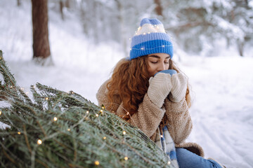 Young woman sits on log and drinks hot drink from thermos in snowy forest. A happy woman enjoys winter forest and drinks tea with a Christmas tree outdoors. Concept of celebration and relaxation.