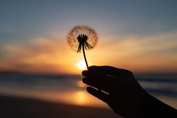Fototapeta premium Silhouette hand holds a dandelion against a setting sun over ocean. Serene and tranquil