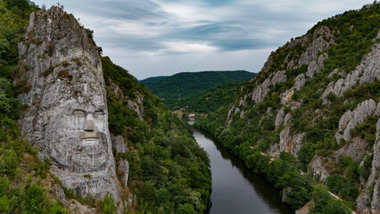 Decebalus Rex Overlooking the Danube's Dramatic Gorges. An expansive aerial or elevated view capturing the majestic Decebalus Rex rock sculpture on the left, carved into a colossal cliff face.
