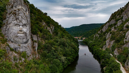 Decebalus Rex Overlooking the Danube's Dramatic Gorges. An expansive aerial or elevated view capturing the majestic Decebalus Rex rock sculpture on the left, carved into a colossal cliff face.