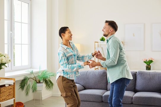 Portrait of happy smiling young couple having fun and dancing at home. Cheerful man and woman enjoying time together standing in living room. Love, people and relationships concept.