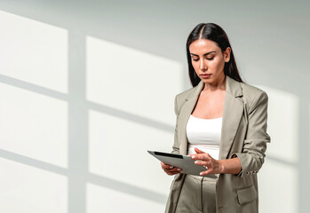 Stylish businesswoman in beige suit using tablet device, realistic style, standing against white wall with window shadow. Concept of modern businesswoman