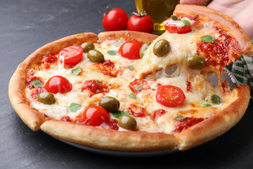 Woman taking piece of delicious pizza with cheese, tomatoes and olives at dark grey table, closeup